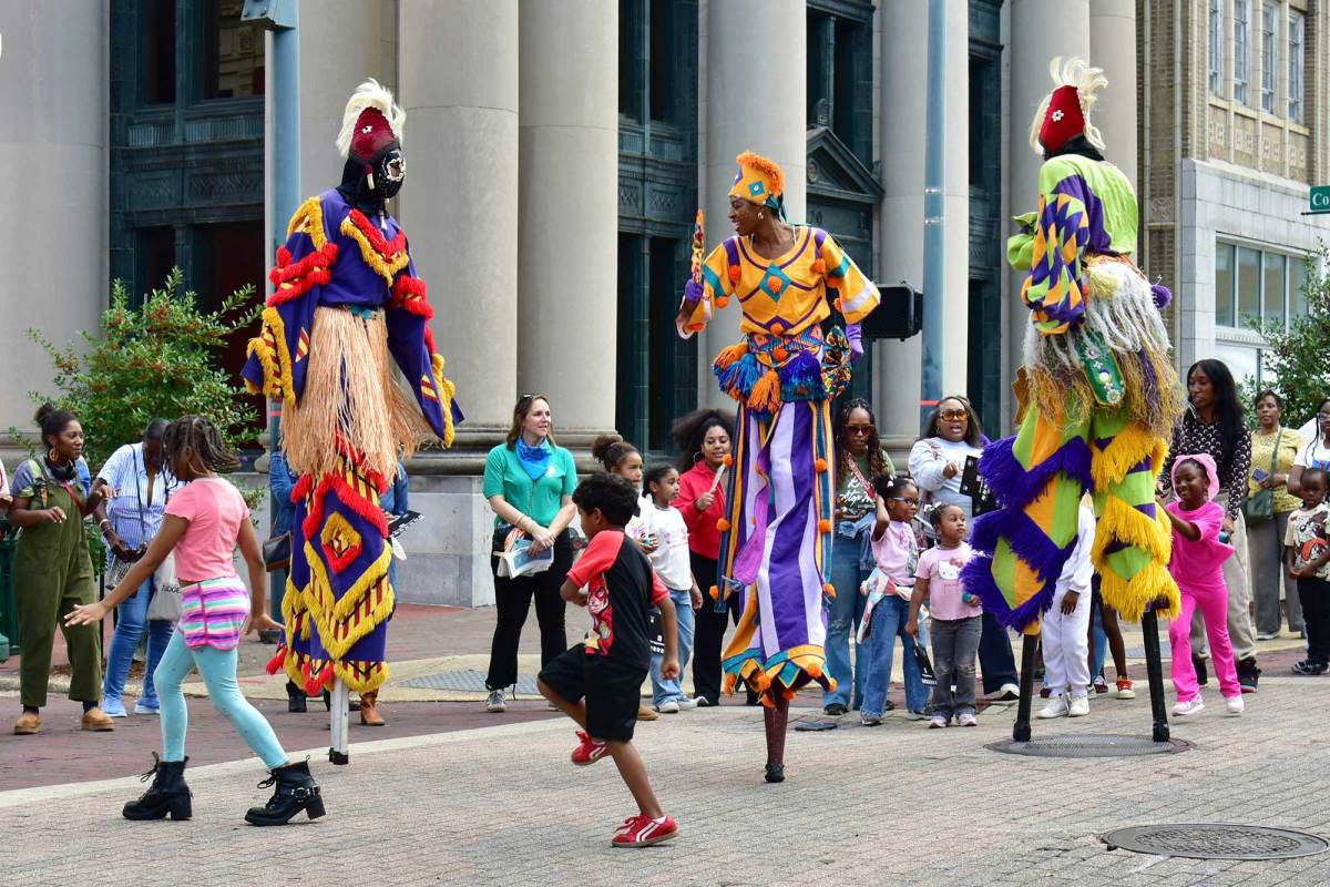 Shaka Zulu dancers perform in colorful outfits and standing on tall stilts as crowds watch on and children dance around them in the streets of downtown Jackson