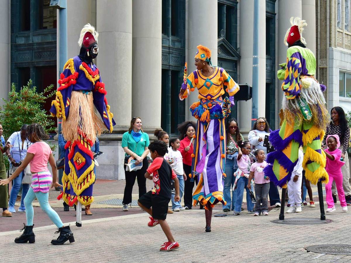 Shaka Zulu dancers perform in colorful outfits and standing on tall stilts as crowds watch on and children dance around them in the streets of downtown Jackson