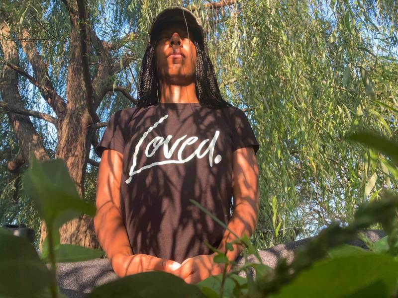 A woman seated in a field with a tree behind her. She is wearing a black shirt that says, “Loved.”