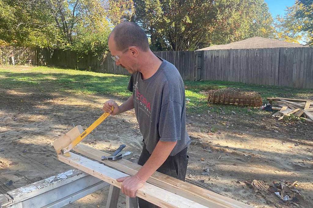 A man cuts wood on a sawhorse in the back yard