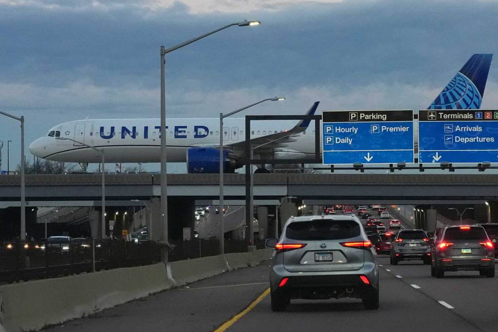 A United Airlines flight lands on a runway. Cars drive under the elevated runway.