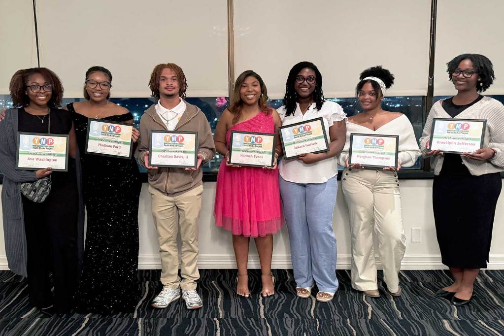Seven teenaged journalists pose in front of a shaded window at night, holding framed certificates with large YMP logos on top