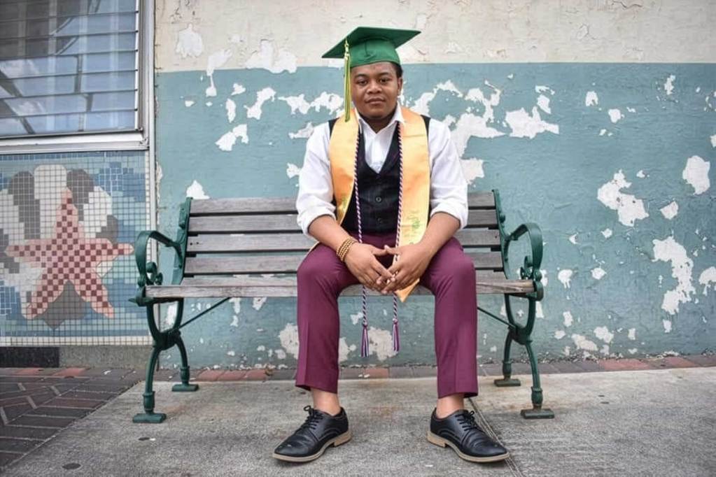 A man in a graduation cap and stole sits on a park bench outside of a blue and white building