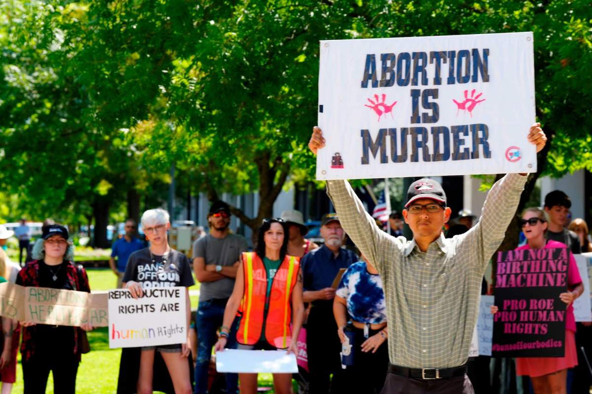 a man standing outside holds up a sign saying "ABORTION IS MURDER." Women standing behind him are holding signs that say "reproductive rights are human rights" and "i am not a birthing machine"