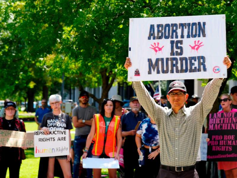 a man standing outside holds up a sign saying "ABORTION IS MURDER." Women standing behind him are holding signs that say "reproductive rights are human rights" and "i am not a birthing machine"