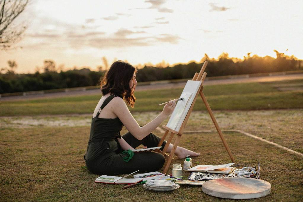 A woman sitting on the ground at sunrise painting on an easel.
