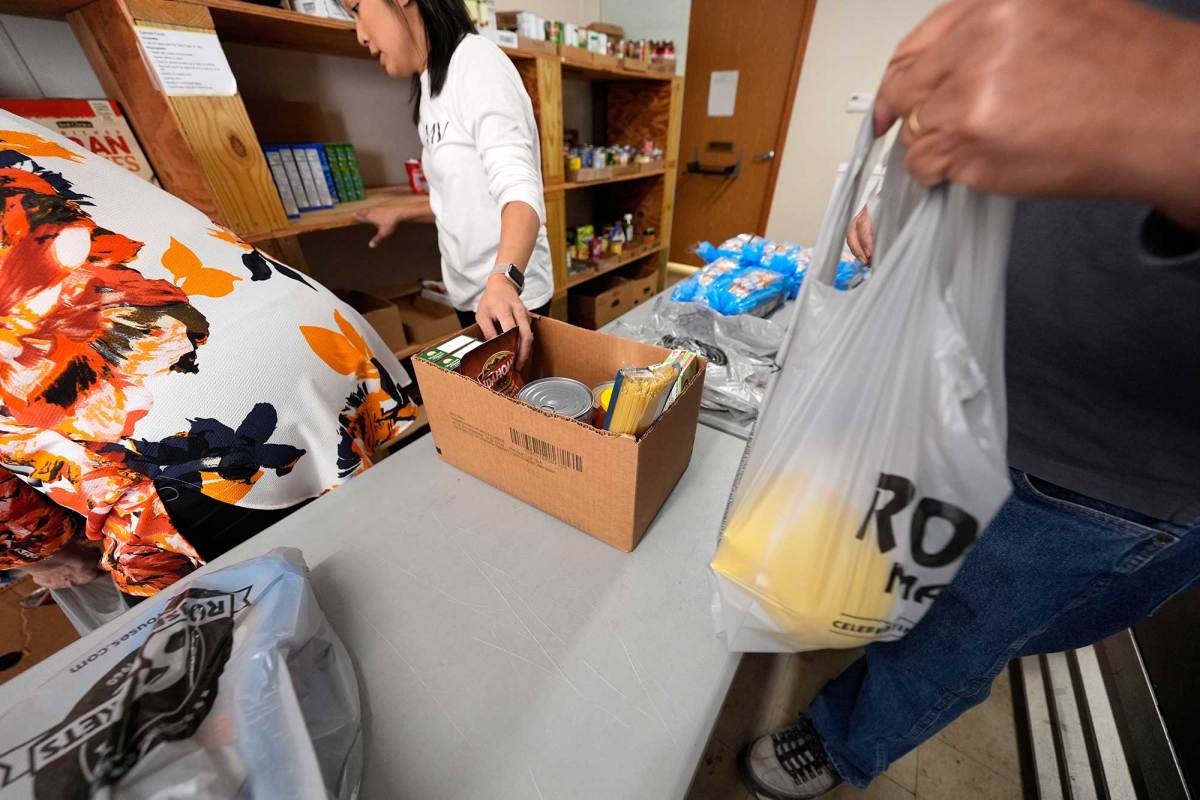Three people at a table handling food supplies, with two women looking at a shelf and one man holding a gray plastic bag. A cardboard box with food supplies is on the table.