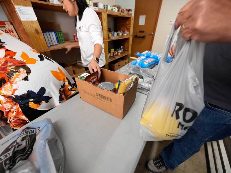 Three people at a table handling food supplies, with two women looking at a shelf and one man holding a gray plastic bag. A cardboard box with food supplies is on the table.