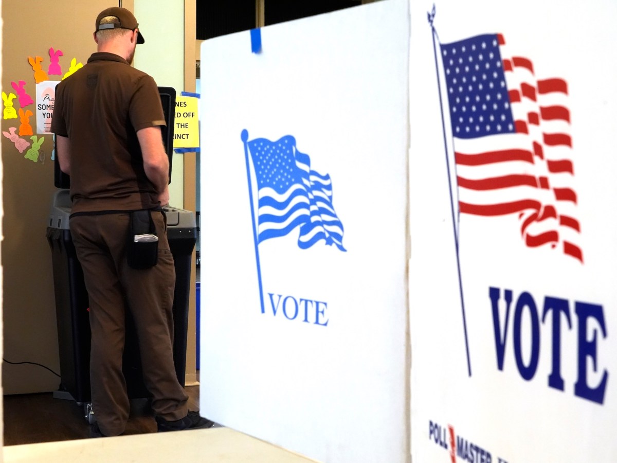 a man in a brown shirt, pants and hat is seen from behind standing at a voting machine. In the foreground there are several "VOTE" boards