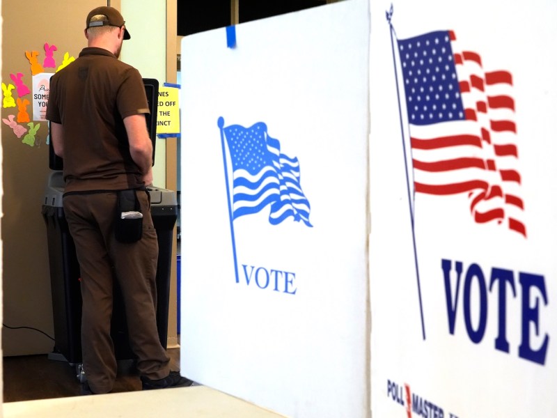 a man in a brown shirt, pants and hat is seen from behind standing at a voting machine. In the foreground there are several "VOTE" boards