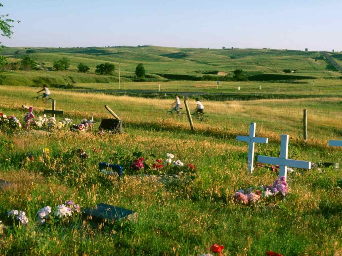 Children riding bikes in the background of an open, grassy field with several white crosses installed in the ground.