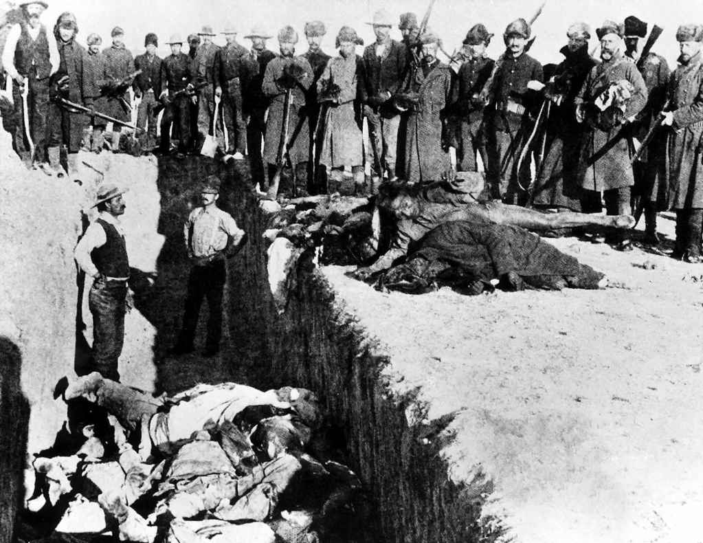 A black-and-white photo of numerous men in uniform standing before an open hole filled with corpses.