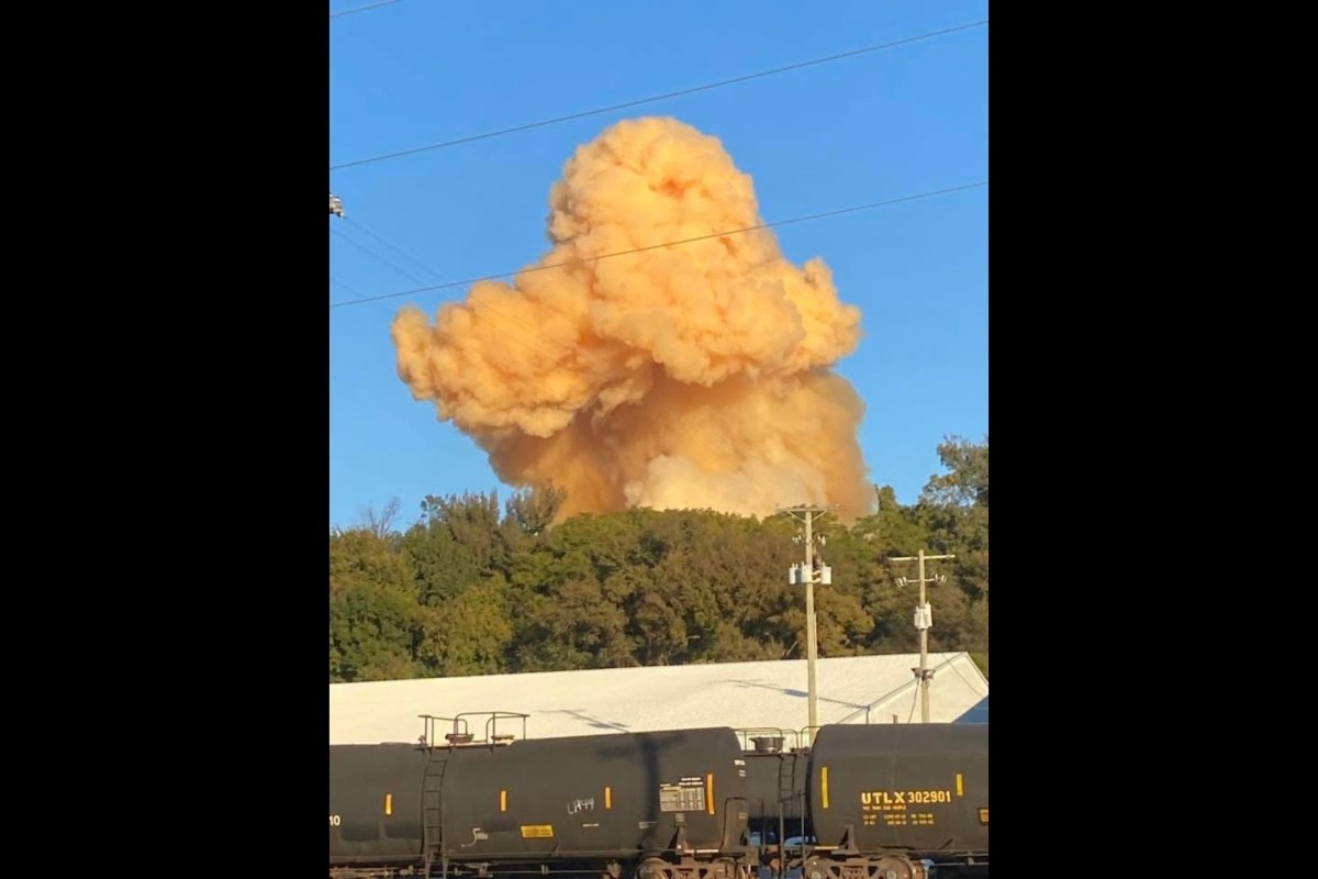A train in the foreground, with trees in the background and an orange explosion rising against a blue sky