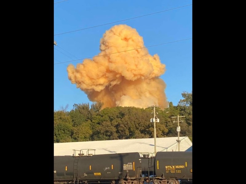 A train in the foreground, with trees in the background and an orange explosion rising against a blue sky