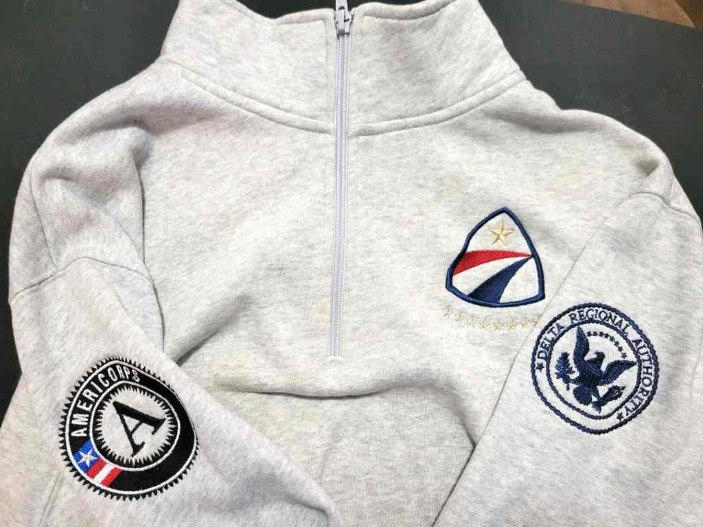 A long-sleeved light grey sweater with the AmeriCorps logo on the right sleeve and the Delta Regional Authority logo on the left sleeve.