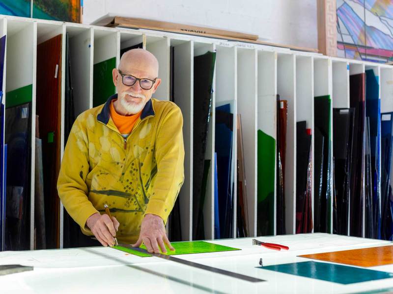 A man in a yellow jacket stands at a lit table top, ready to cut into colorful sheets of glass