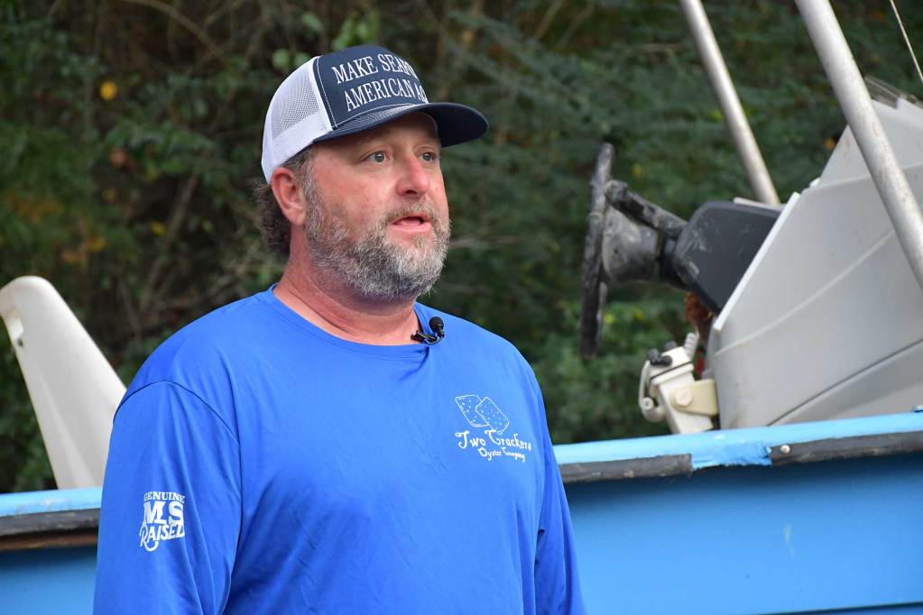 A man in a blue, long-sleeve shirt with a logo “Two Crackers Oyster Company” and a hat that says “Make Seafood American Again” stands in front of a boat.