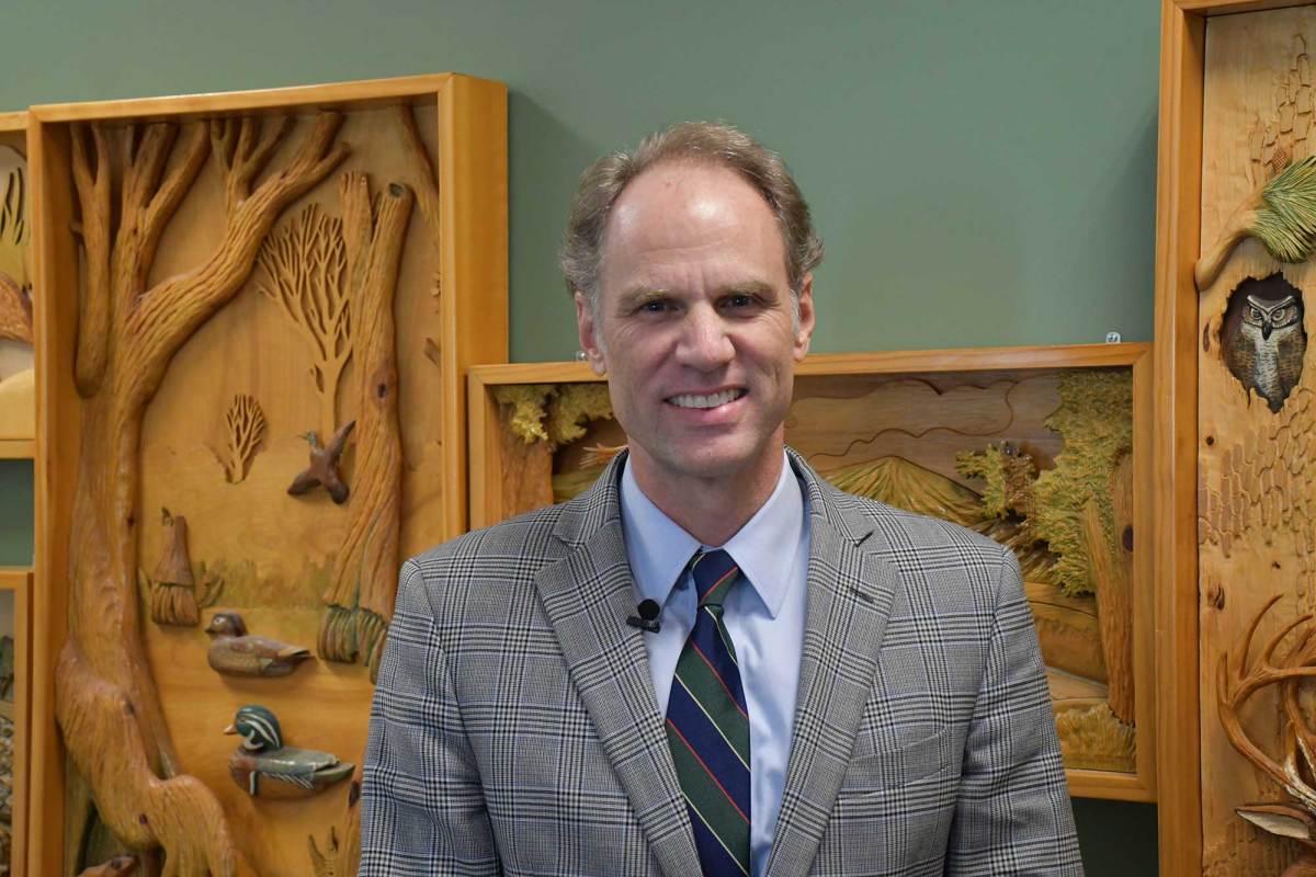 A man wearing a gray plaid suit jacket, blue shirt and striped tie stands indoors in front of carved wooden nature-themed wall art, smiling at the camera.