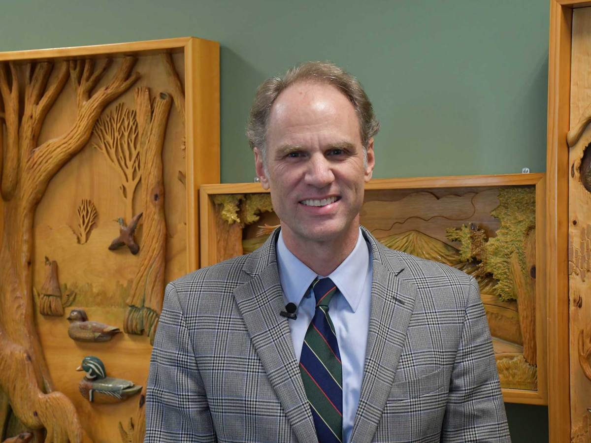 A man wearing a gray plaid suit jacket, blue shirt and striped tie stands indoors in front of carved wooden nature-themed wall art, smiling at the camera.