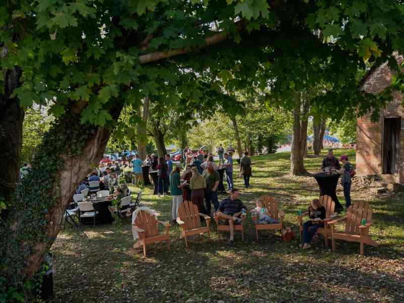 A gathering of people in a park, with some seated in large wooden chairs and several others standing.