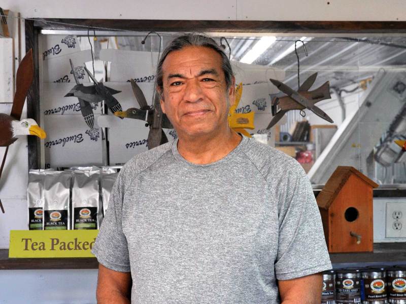 A man in a gray t-shirt and dark gray shorts smiles at the camera, standing in front of product display shelves inside J&D Blueberry Farms shop.