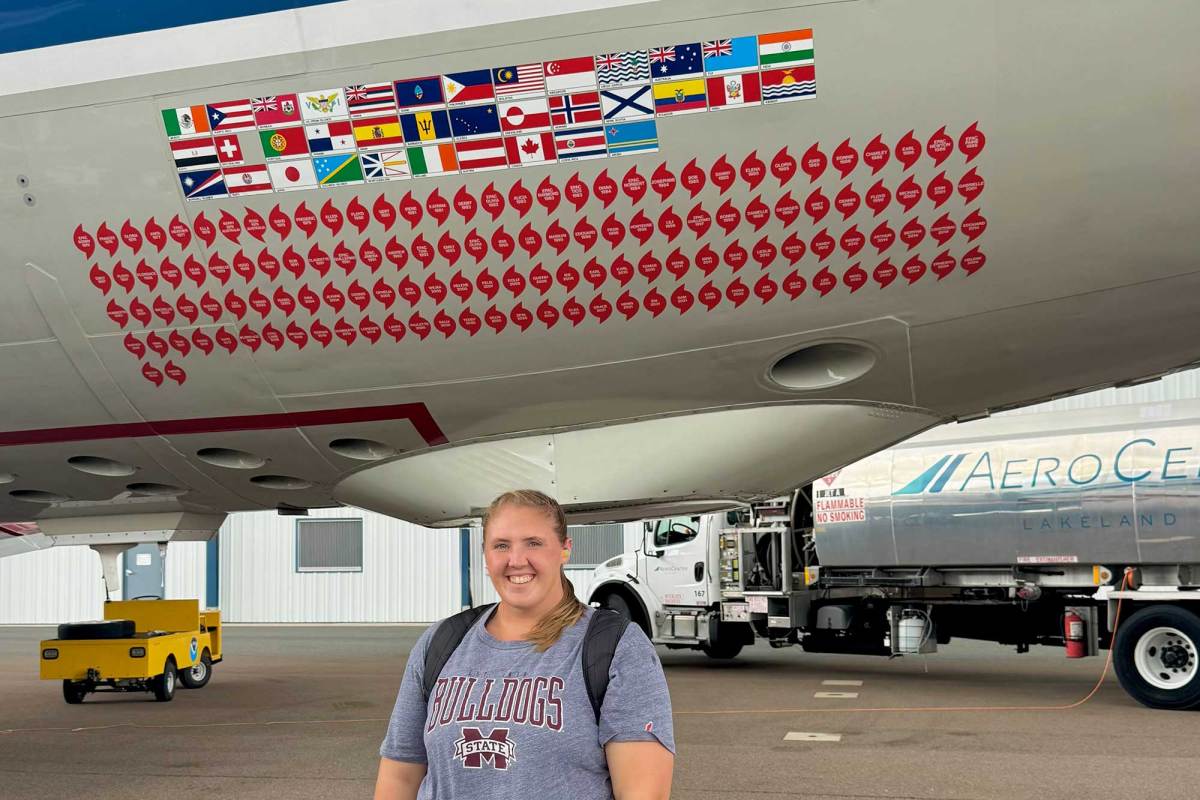 A woman in a Mississippi State tshirt stands on an airport runway in front of a parked plane covered in stickers of flags and hurricane symbols