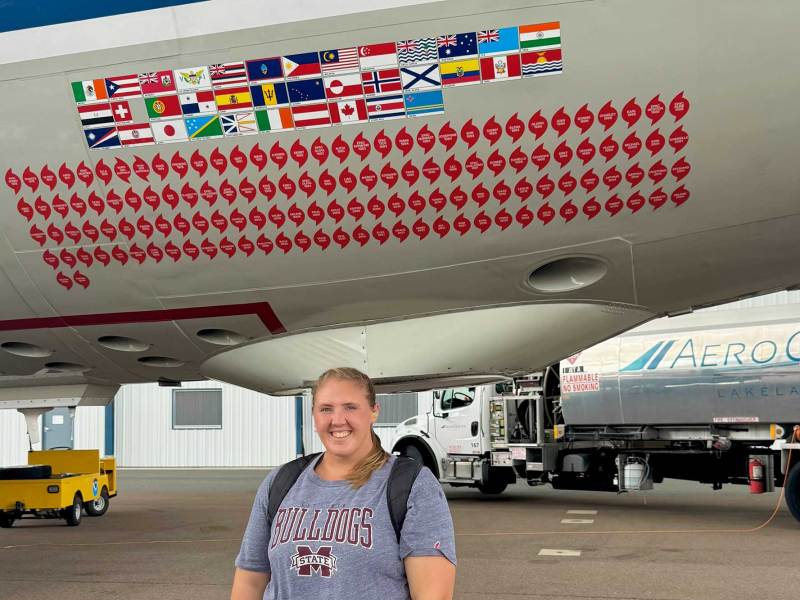 A woman in a Mississippi State tshirt stands on an airport runway in front of a parked plane covered in stickers of flags and hurricane symbols