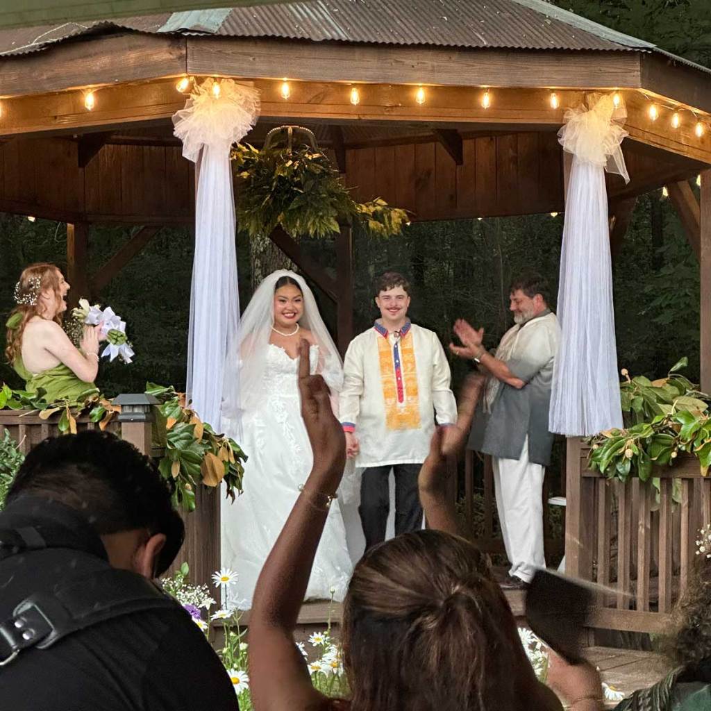 People in wedding dress stand in a lit up gazebo as people clap around them