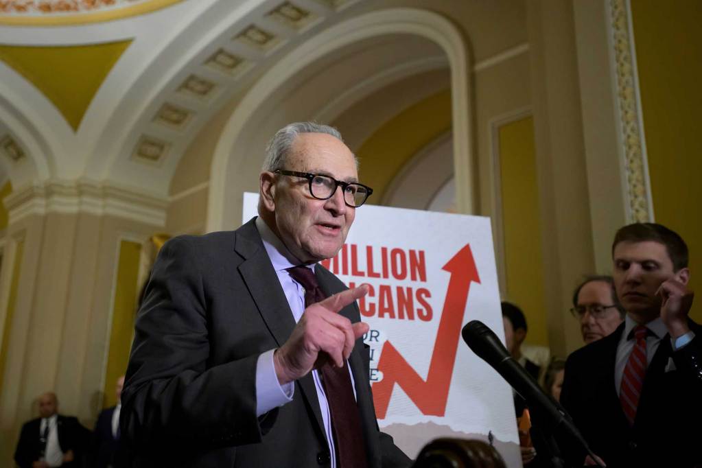 Chuck Schumer speaking at a podium. Behind him is a chart with a red arrow leading up