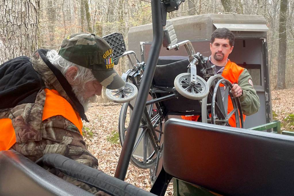 Two men in hunting camo and orange high-viz vests work to get a wheelchair into an offroad vehicle