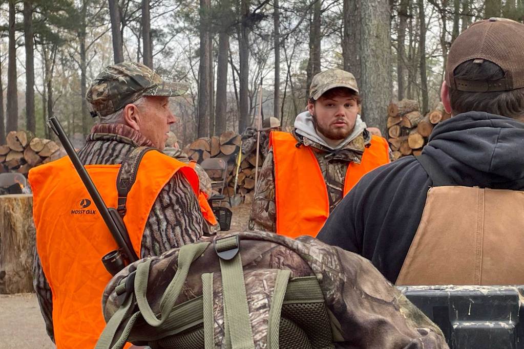 Three people in hunter's camo and orange high-viz vests stand outside with hunting rifles