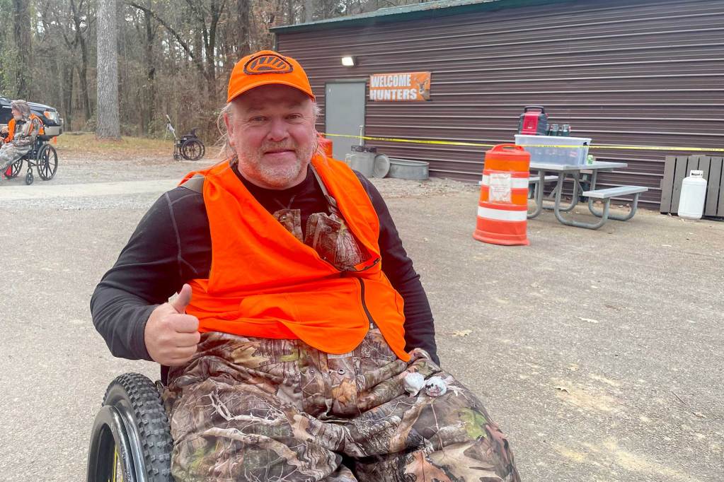 A person in hunting camo and an orange high-viz vest poses thumbs-up in his wheelchair outside of a hunting camp
