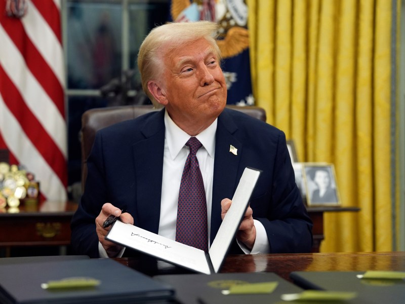 President Donald Trump smiling at his desk in the Oval Office as he closes a folder.