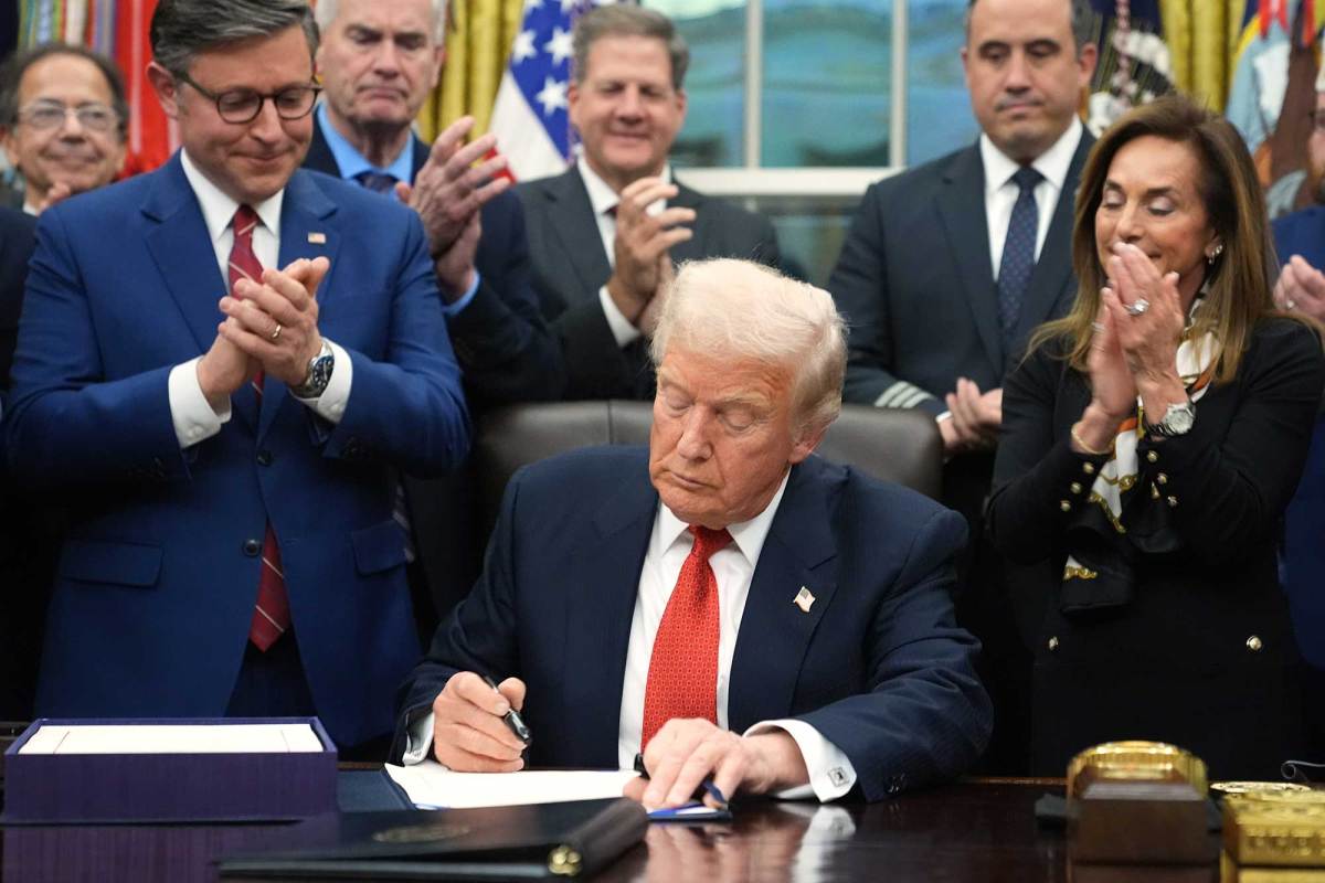 Donald Trump sits at his desk in the Oval Office and signs something as people clap behind him