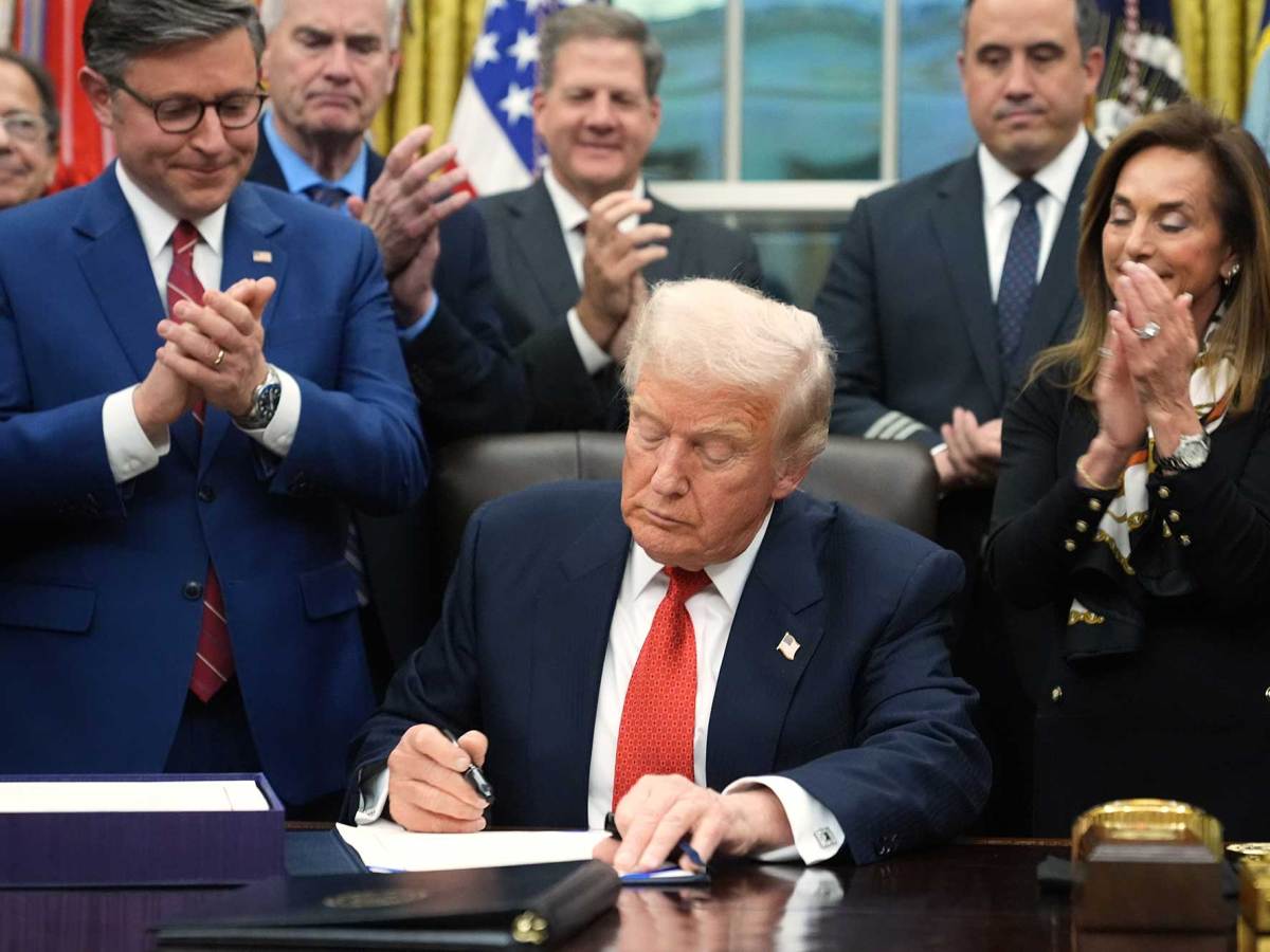 Donald Trump sits at his desk in the Oval Office and signs something as people clap behind him