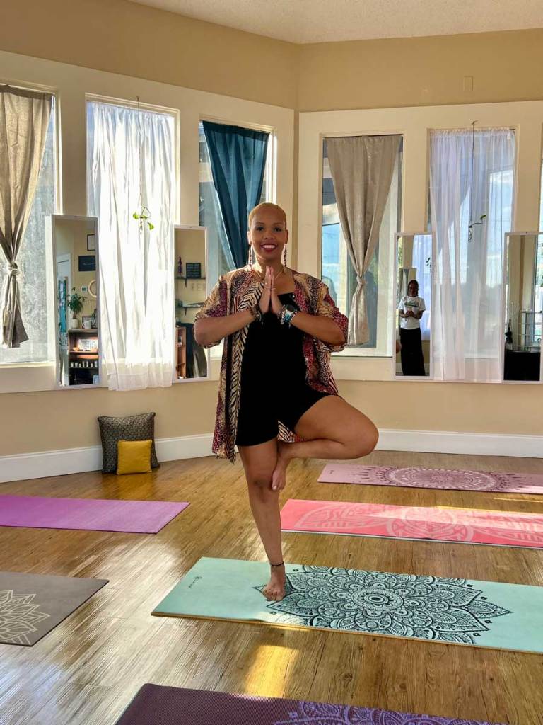 A woman smiling and balancing on one leg in a sunlit yoga studio.