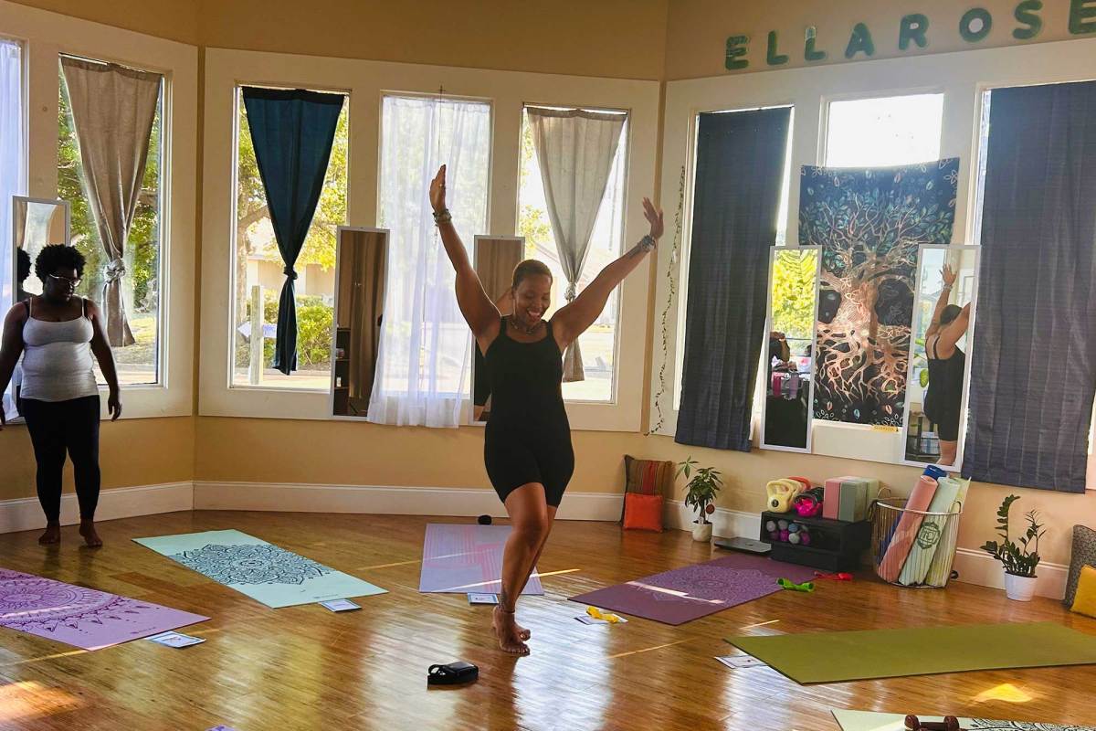 A woman smiling with her hands in the air. Yoga mats are laid out around her in a sunlit room.