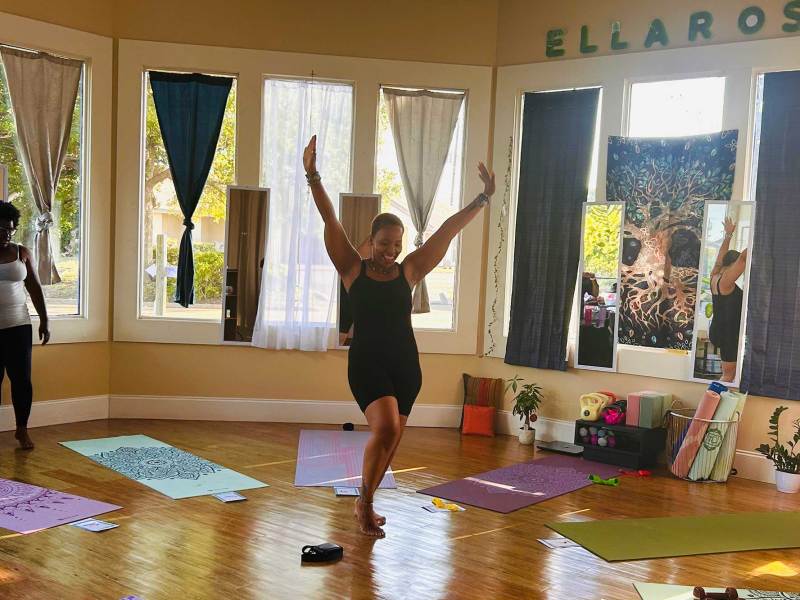 A woman smiling with her hands in the air. Yoga mats are laid out around her in a sunlit room.