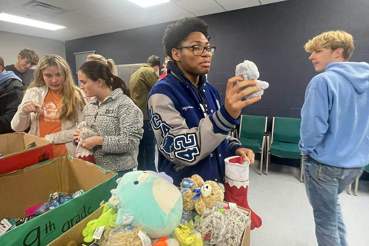 A kid in a room full of others, holding up a plush toy while stuffing stockings