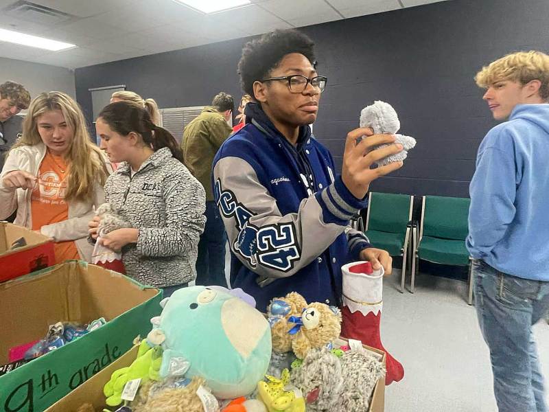 A kid in a room full of others, holding up a plush toy while stuffing stockings