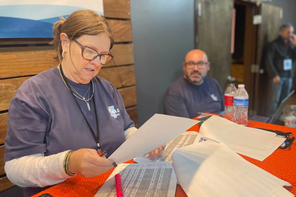 Two people in grey "Halo of Hope' shirts sit behind a red table and read through piles of spreadsheets
