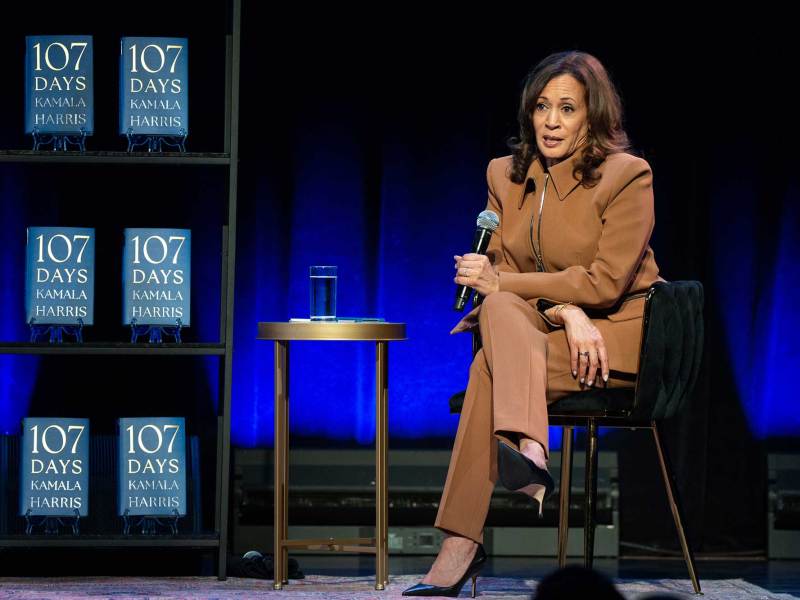Kamala Harris sits on a stage and speaks next to a display of her book '107 Days'