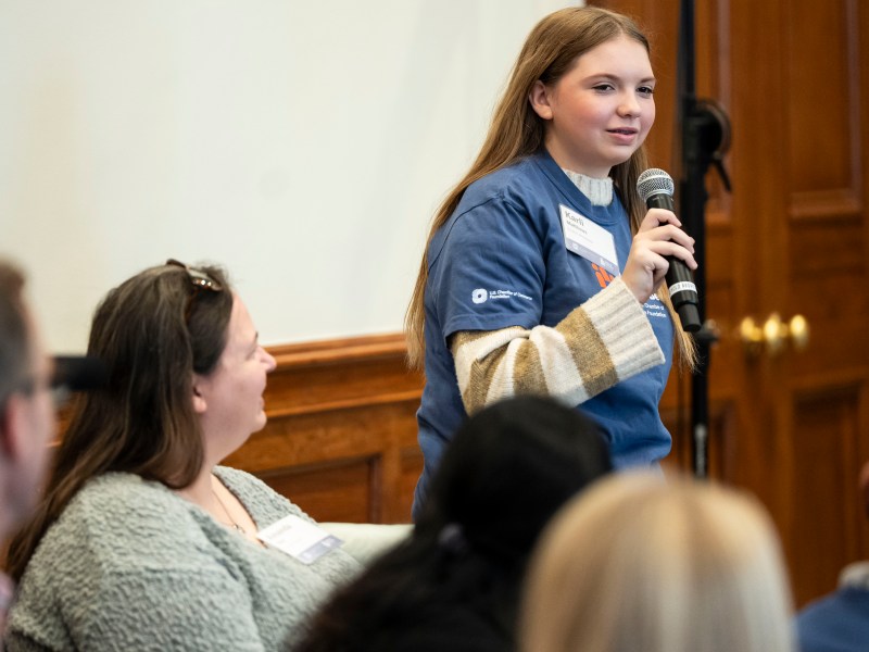 A young woman holds up a mic and speaks to a room at a Civics Bee