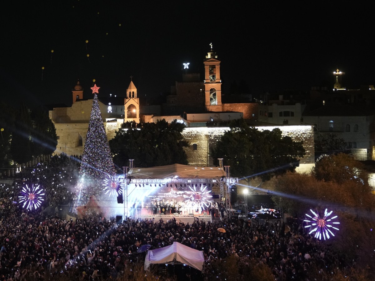 A large gathering of people in a city square around a large Christmas tree and a stage.