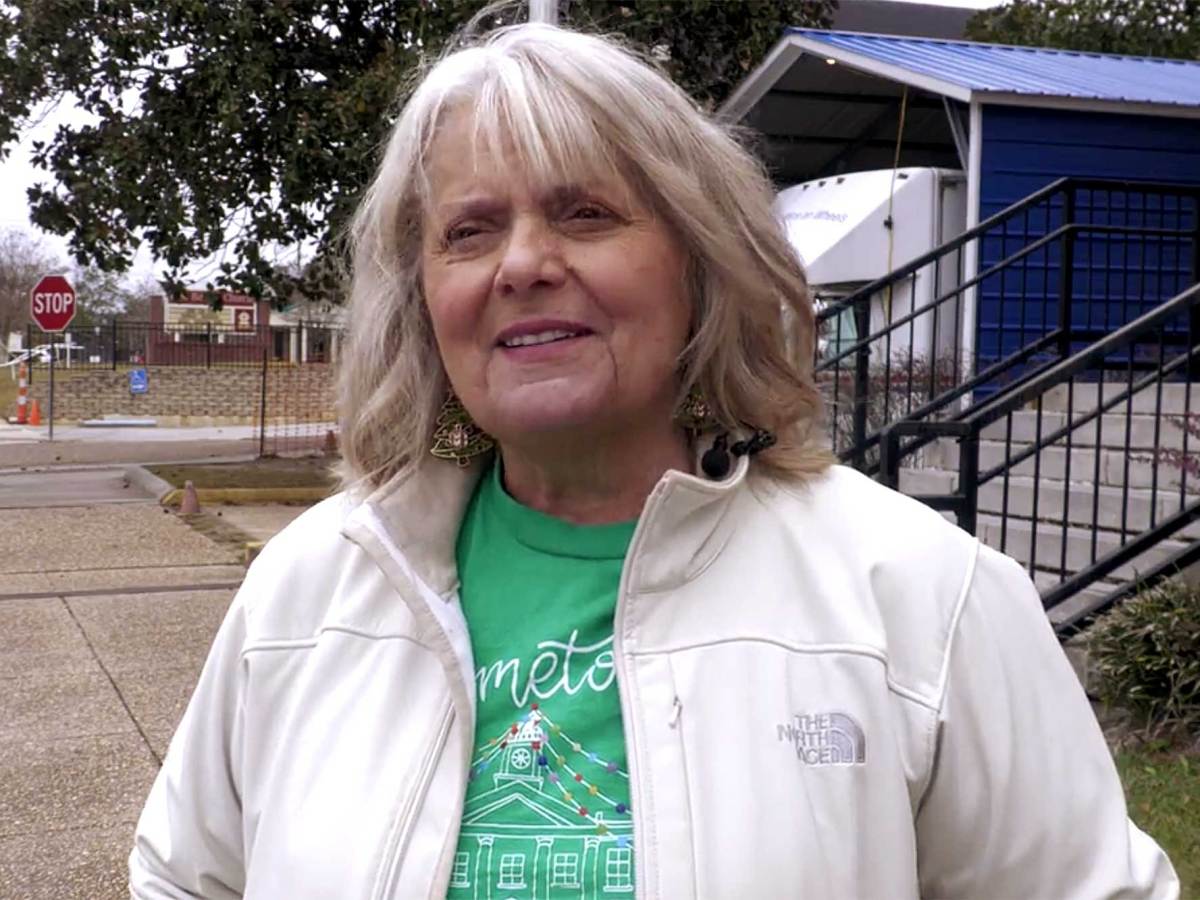 A woman with shoulder-length white hair wearing a white jacket and greet shirt stands on a sidewalk in front on a brick building.