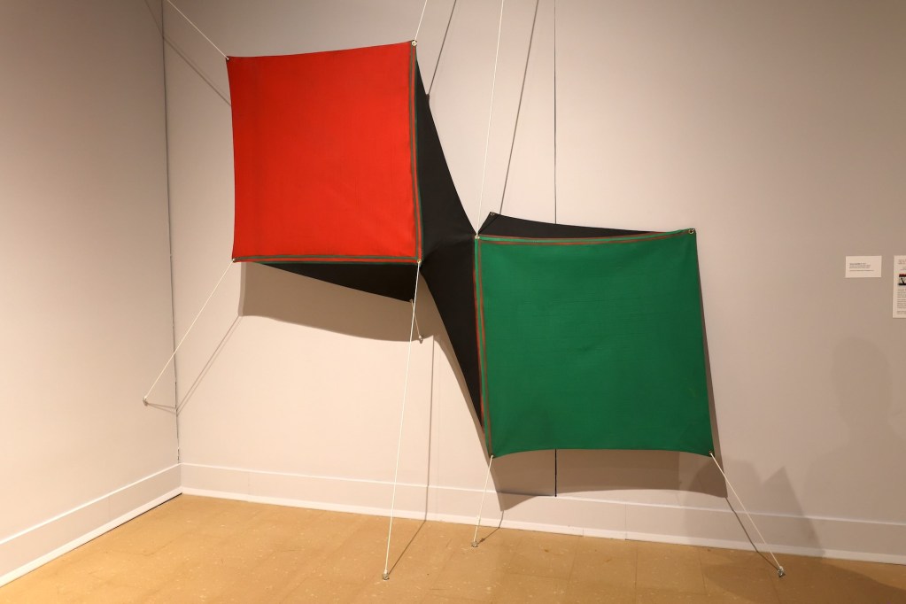 Two 3D pieces, a red cube and a green cube, hang suspended by string