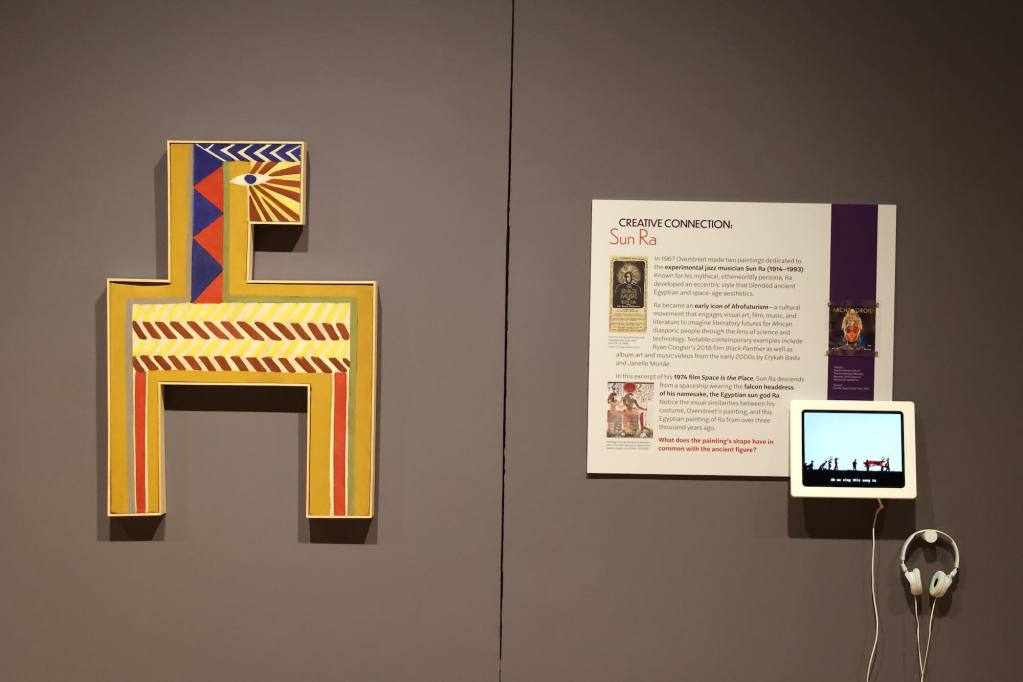An angular art piece painted in an Egyptian style hangs on the gallery wall