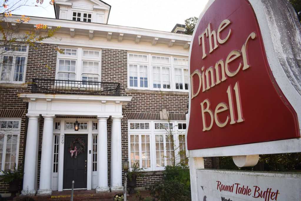 A red bell-shaped sign outside a two-story brick building that reads, “The Dinner Bell.”