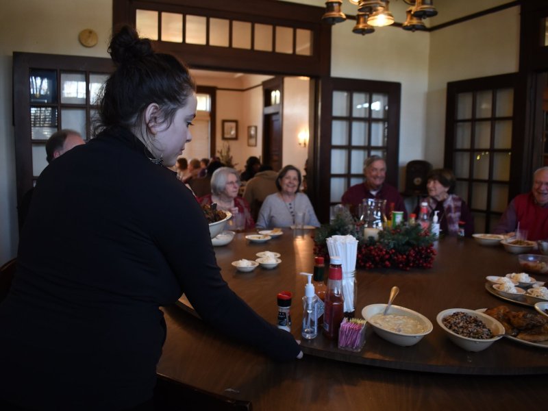 A woman wearing black adjusts a round table around which several people are seated.