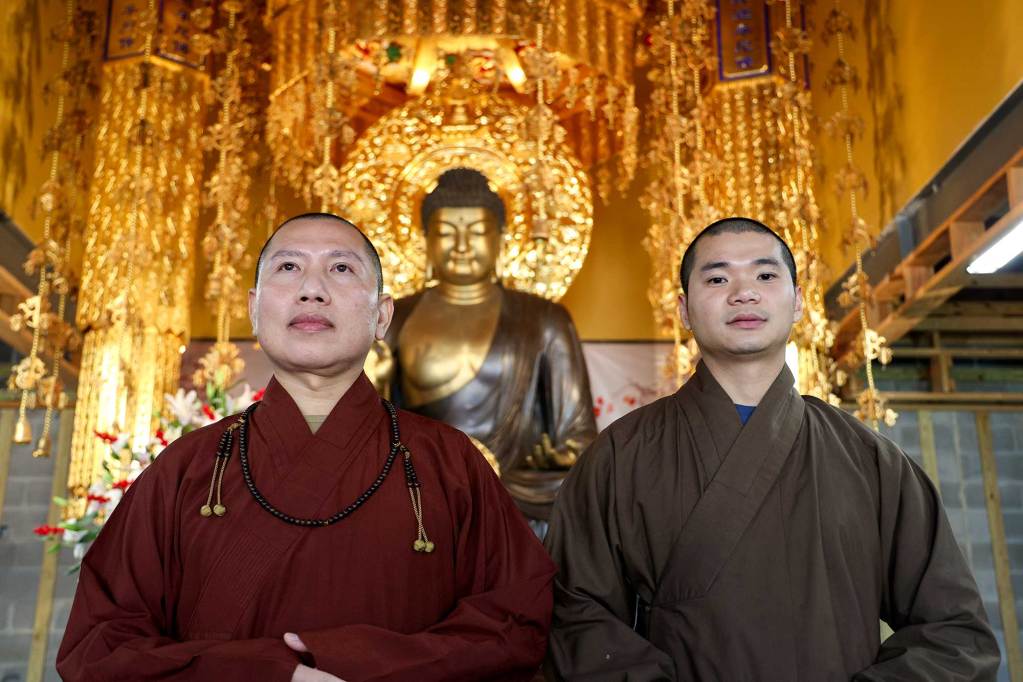 A closeup of a two monks standing in front of a Buddha statue inside a golden decorated temple